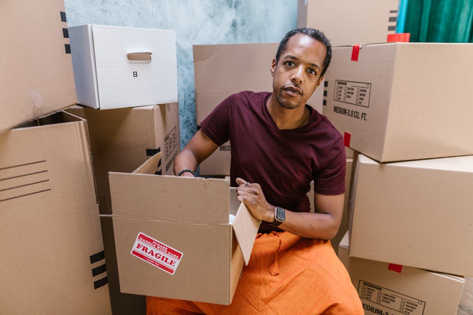 A man with dark hair and medium skin tone is sitting among numerous cardboard moving boxes inside a room, surrounded by packed and partially unpacked cartons. He is wearing a maroon T-shirt, orange pa