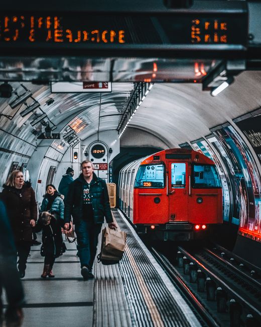 This image depicts a busy underground station platform with several passengers walking alongside the train tracks. In the foreground, a man is carrying a large brown paper bag, while other travelers, including a woman and a child, are walking nearby with personal belongings. The platform is illuminated by artificial lighting, and the curved ceiling features digital displays showing train times and destinations. A red train with illuminated windows is approaching or stationary at the platform, with its doors visible. The scene is inside a modern urban underground station, highlighting the movement and logistics involved in home relocation or furniture transport through the underground rail network. The image reflects the type of logistical considerations involved in professional removals services, such as those offered by Man with Van Balham, during house removals and packing and moving operations, especially in busy city environments.