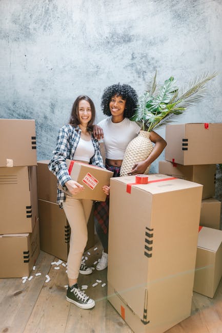 Two women are standing inside a room with a textured, light grey wall, surrounded by several large cardboard boxes sealed with red and black packing tape. One woman, wearing a plaid shirt and beige pants, holds a medium-sized box with a red 'Fragile' sticker and is smiling at the camera. The other woman, dressed in a grey t-shirt and checkered trousers, is smiling and holding a large woven basket featuring a leafy green plant. The boxes are arranged in a semi-circle around them, some stacked on the floor and others placed against the wall. The scene is illuminated with soft, natural lighting, suggesting a home relocation process involving packing and preparing household items for transportation. This image reflects the careful handling of furniture and packing materials typical of a professional removals service such as Man with Van Balham, supporting efficient furniture transport and moving logistics during house removals or home relocation.